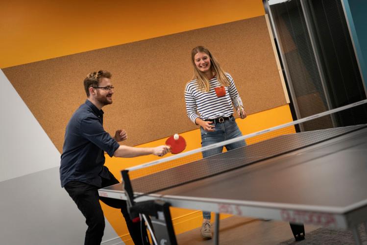 Two colleagues enjoying a doubles game of table tennis in front of a yellow background. One is about to return the ball and they are both smiling.
